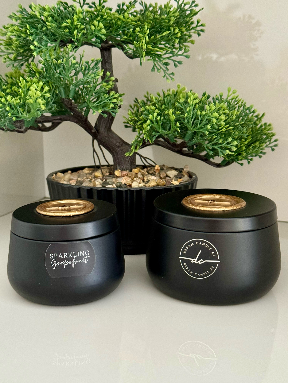 Two black candles with gold lids on a white surface, with a bonsai tree in the background.