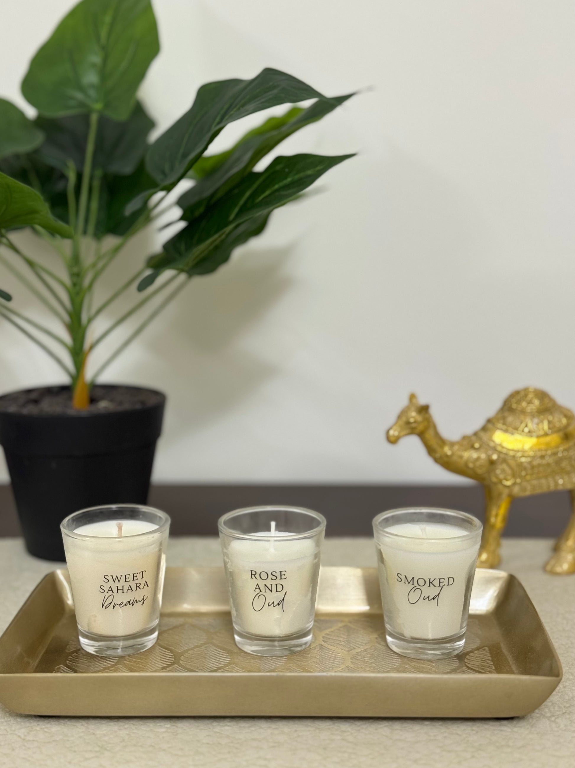 Three small glass candles on a gold tray with a plant and decorative camel in the background.