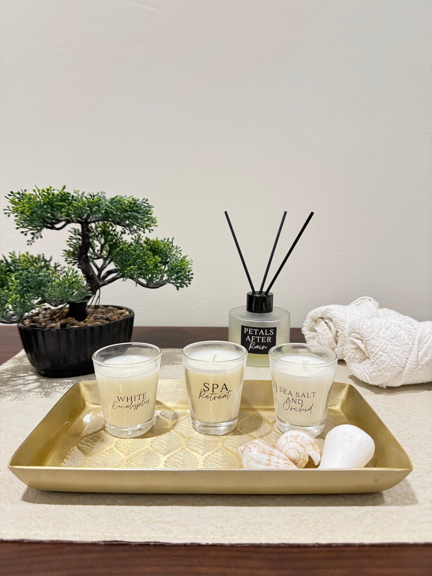 Candle set with 'White Petals' and 'Spa' scents on a gold tray with a plant and towel in the background.