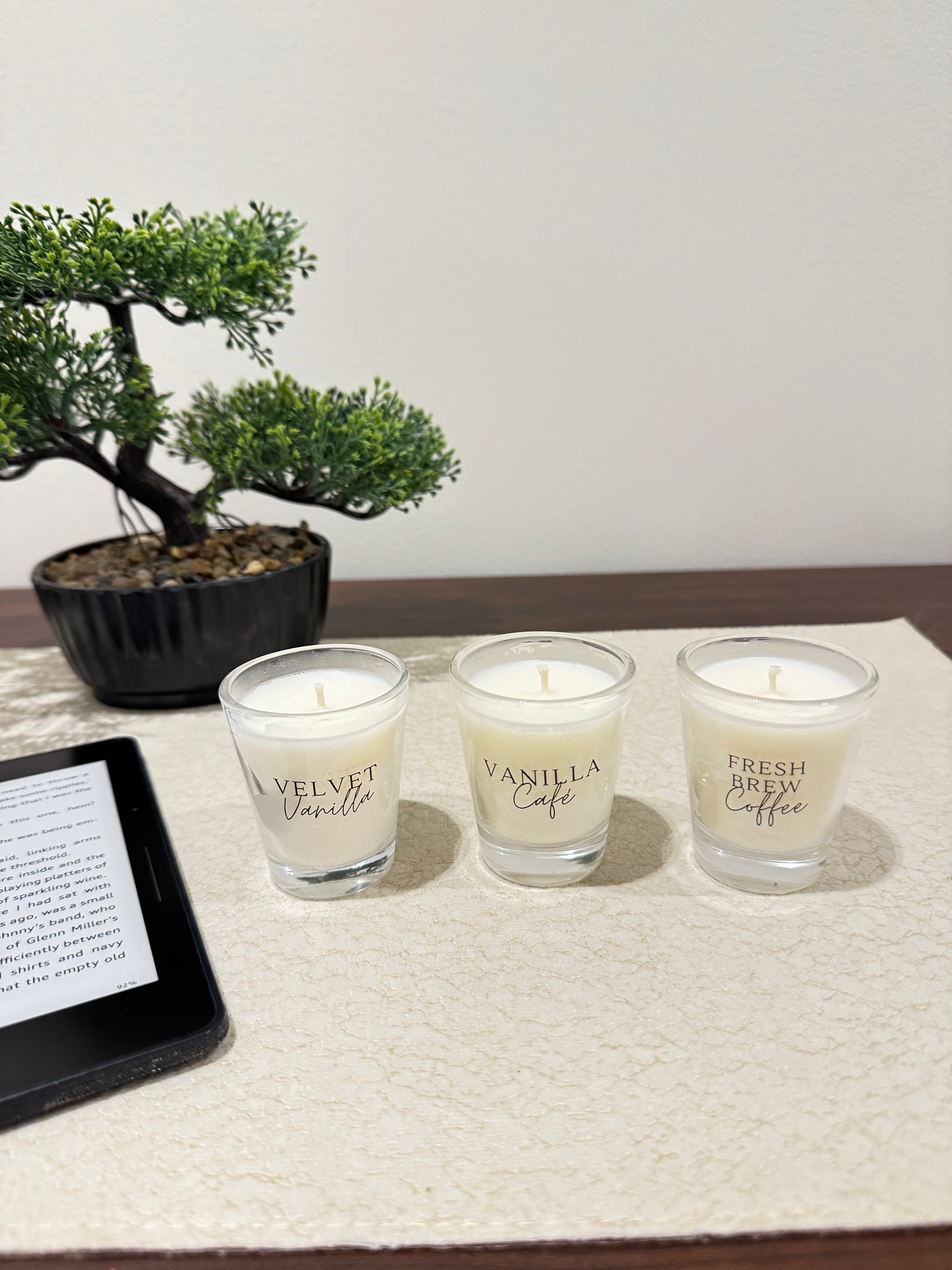 Three scented candles labeled 'Velvet Vanilla', 'Vanilla Cafe', and 'Fresh Brew Coffee' on a table with a tablet and bonsai tree.
